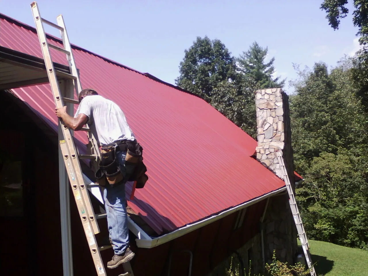 Roofer on a ladder beside a red metal roof and stone chimney — Soffit & Fascia Repair by Roofing Brothers in La Jose